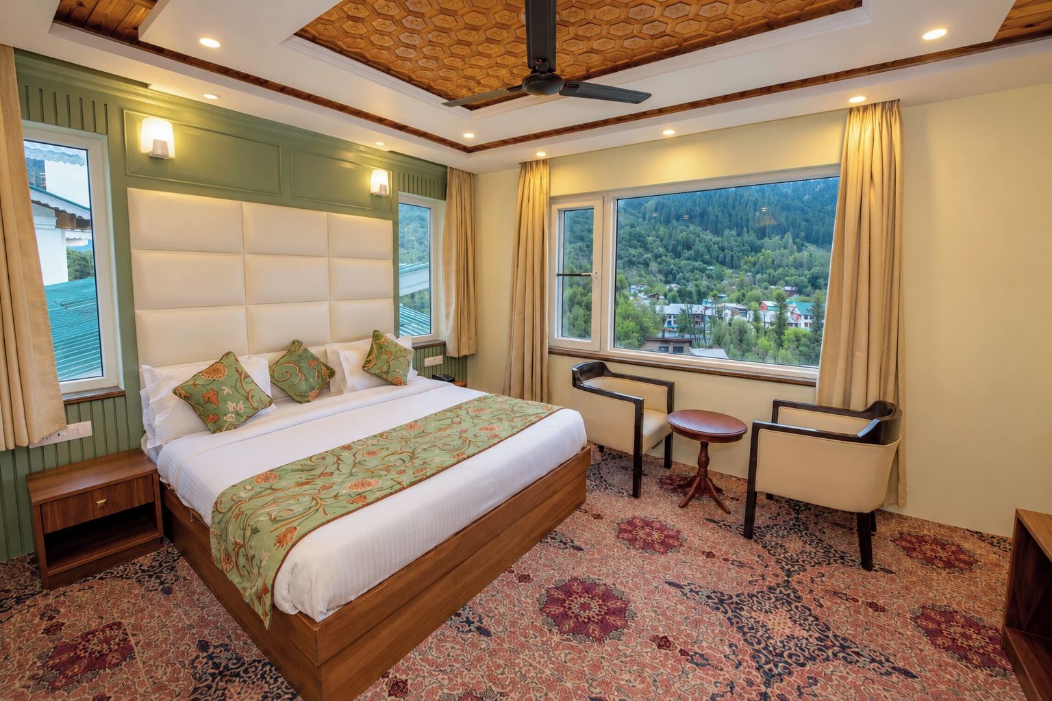 A hotel room with a large bed, a small table, a chair, and a view of a forest through the window.