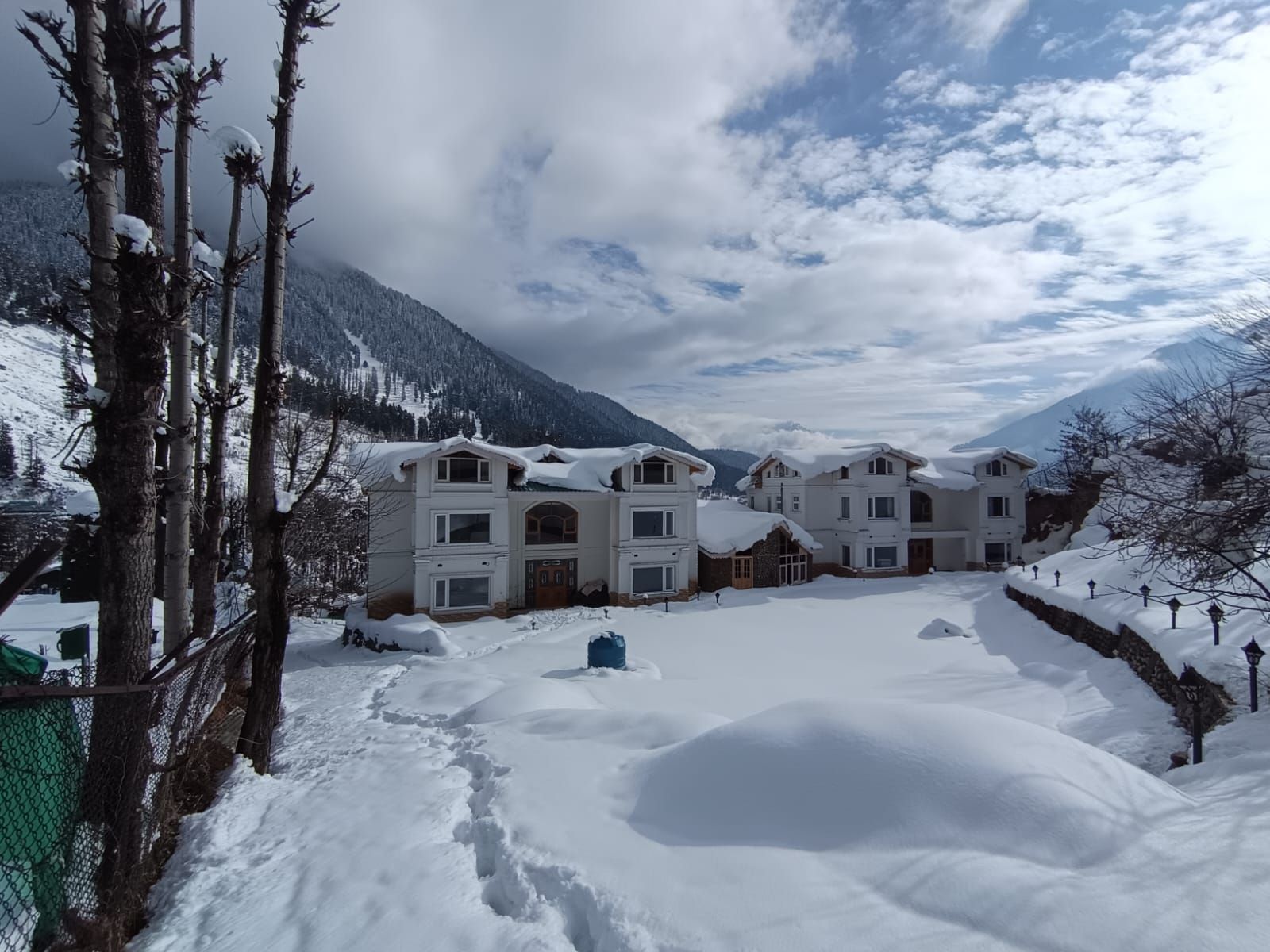 A snow-covered residential area with multiple houses, a fenced pathway, and a mountainous backdrop.