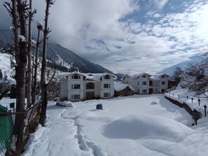 Un barrio residencial cubierto de nieve, con varias casas, un sendero cercado y montañas al fondo.