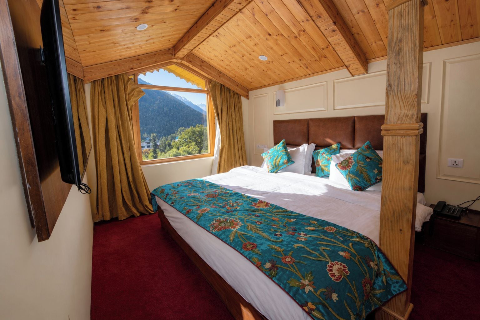 A bedroom with a wooden ceiling, a large bed with floral bedding, a view of mountains through a window, and a red carpet.