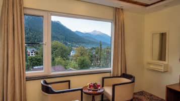 A hotel room with a large window offering a mountain view, two chairs, a small table with a drink, and a mirror on the wall.