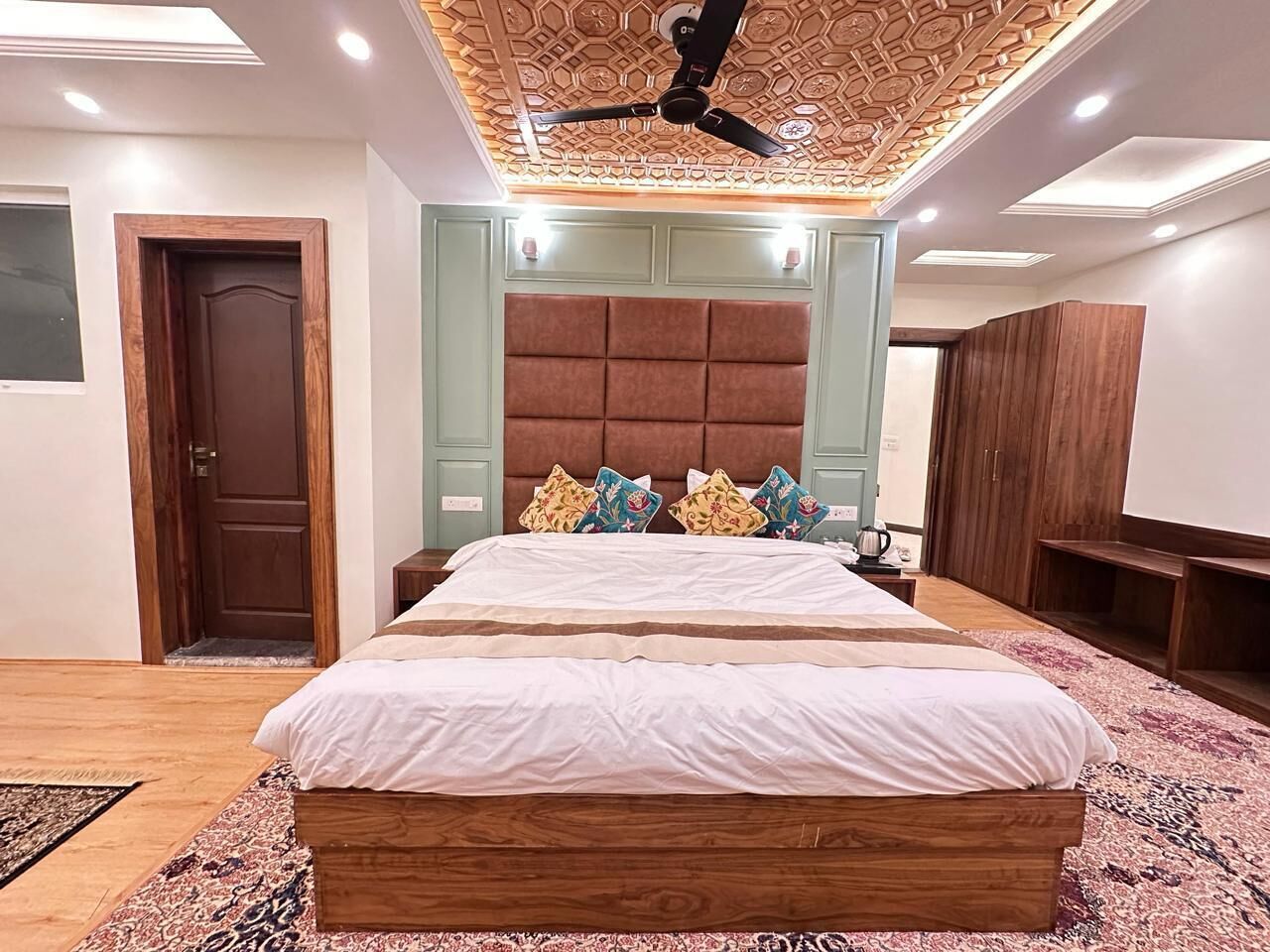 Un dormitorio con cama de madera, ventilador de techo, cabecera acolchada grande y alfombra estampada.