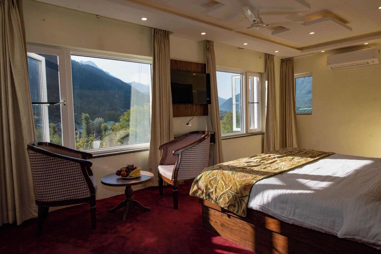 A hotel room with a bed, two chairs, a small table with fruit, and a view of mountains through large windows.