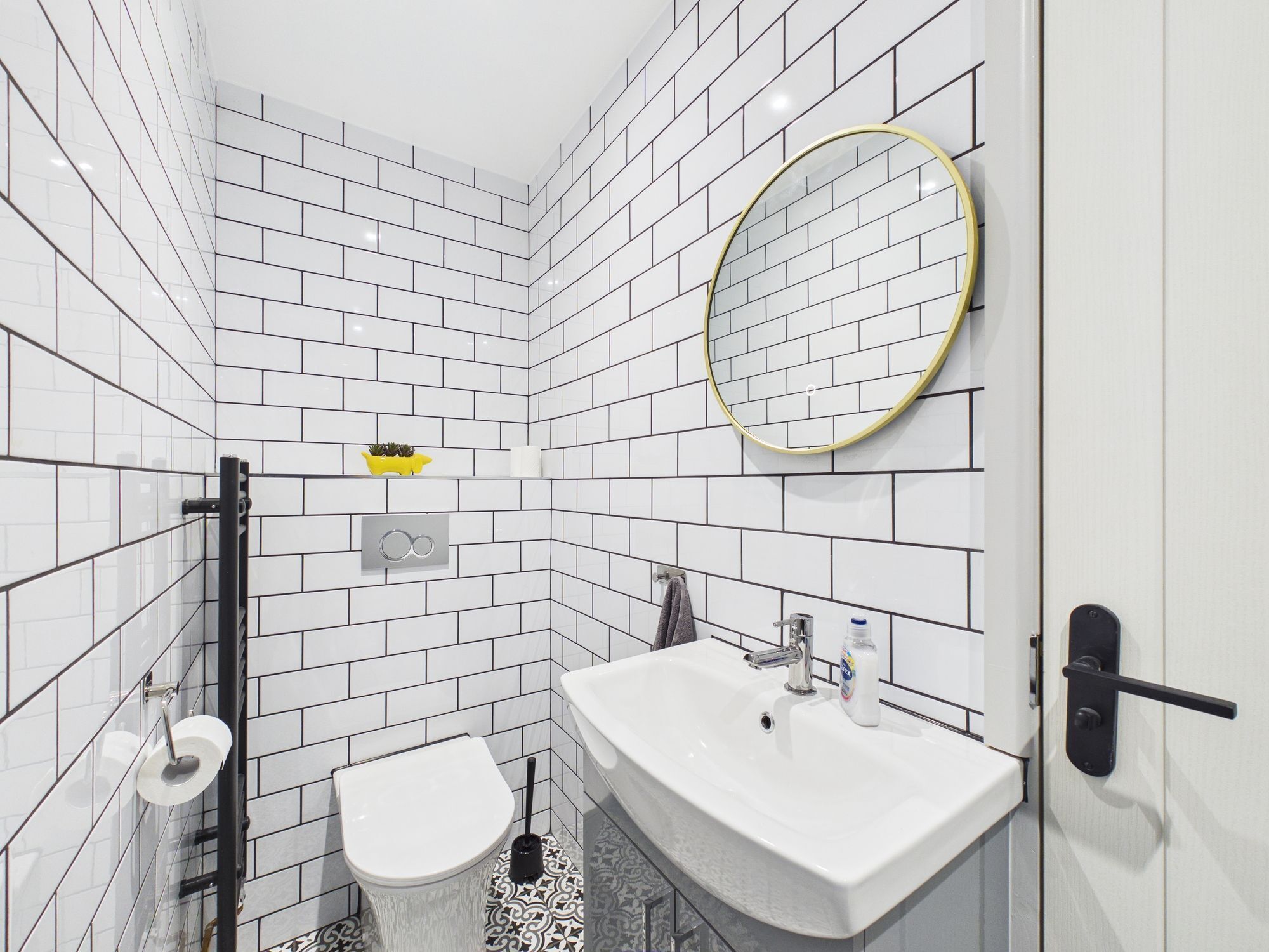 Un baño moderno con azulejos blancos tipo metro, un lavamanos blanco, un inodoro, un espejo redondo y un estante pequeño con un objeto amarillo.
