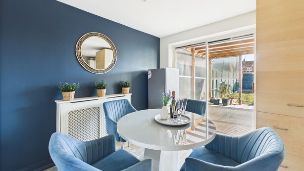 A modern dining area with a round table, blue upholstered chairs, a white cabinet, and a large window with a view of the outdoors.