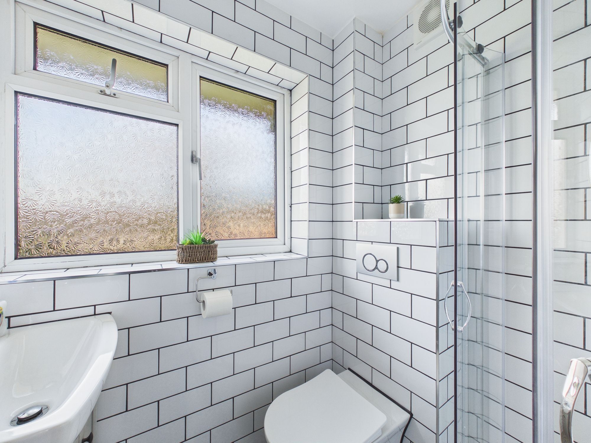 Ein modernes Badezimmer mit weißen, im Fischgrätmuster angeordneten Fliesen, einer Glasdusche, einer Toilette und einem Fenster mit Milchglas.