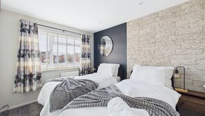 A modern bedroom with two beds, a large window with blinds, a stone wall, and a round mirror.