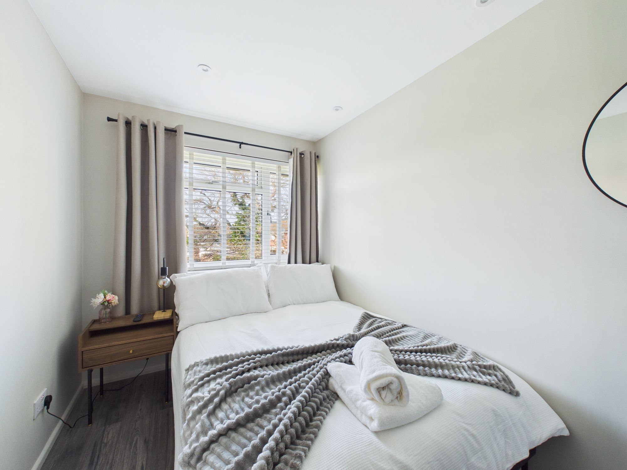A neatly made bed with a rolled towel, a bedside table with a lamp, and a window with curtains.