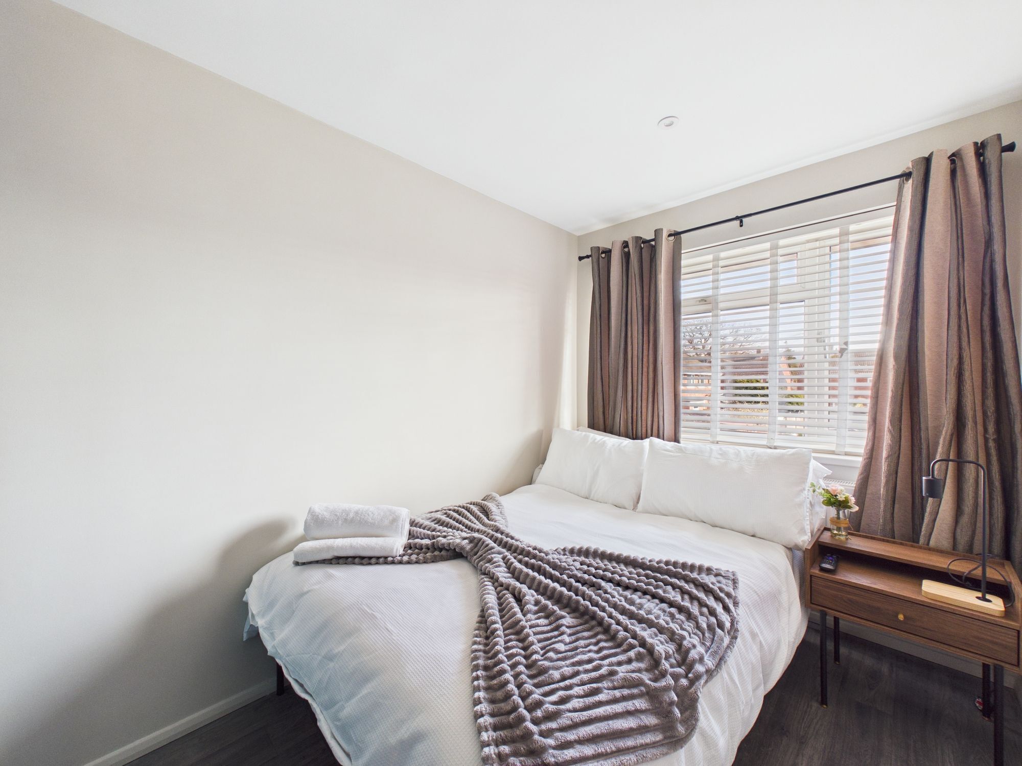 A neatly made bed with a blanket, a bedside table with a lamp, and a window with curtains.