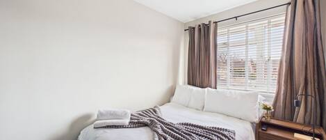 A neatly made bed with a blanket, a bedside table with a lamp, and a window with curtains.