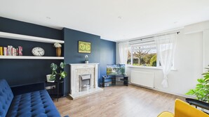 A living room with a blue sofa, a yellow armchair, a fireplace, a TV, and a bookshelf with books and a clock.