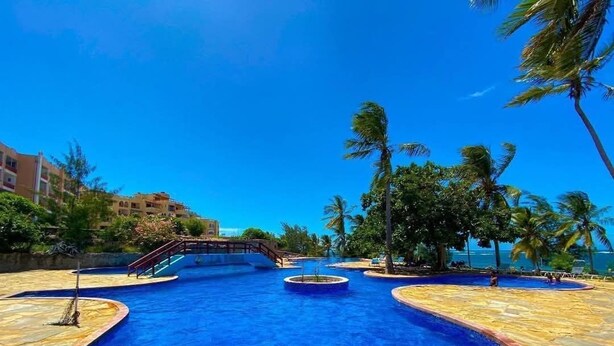 Una piscina grande y cristalina de color azul con un deck de piedra, rodeada de palmeras y un puente.