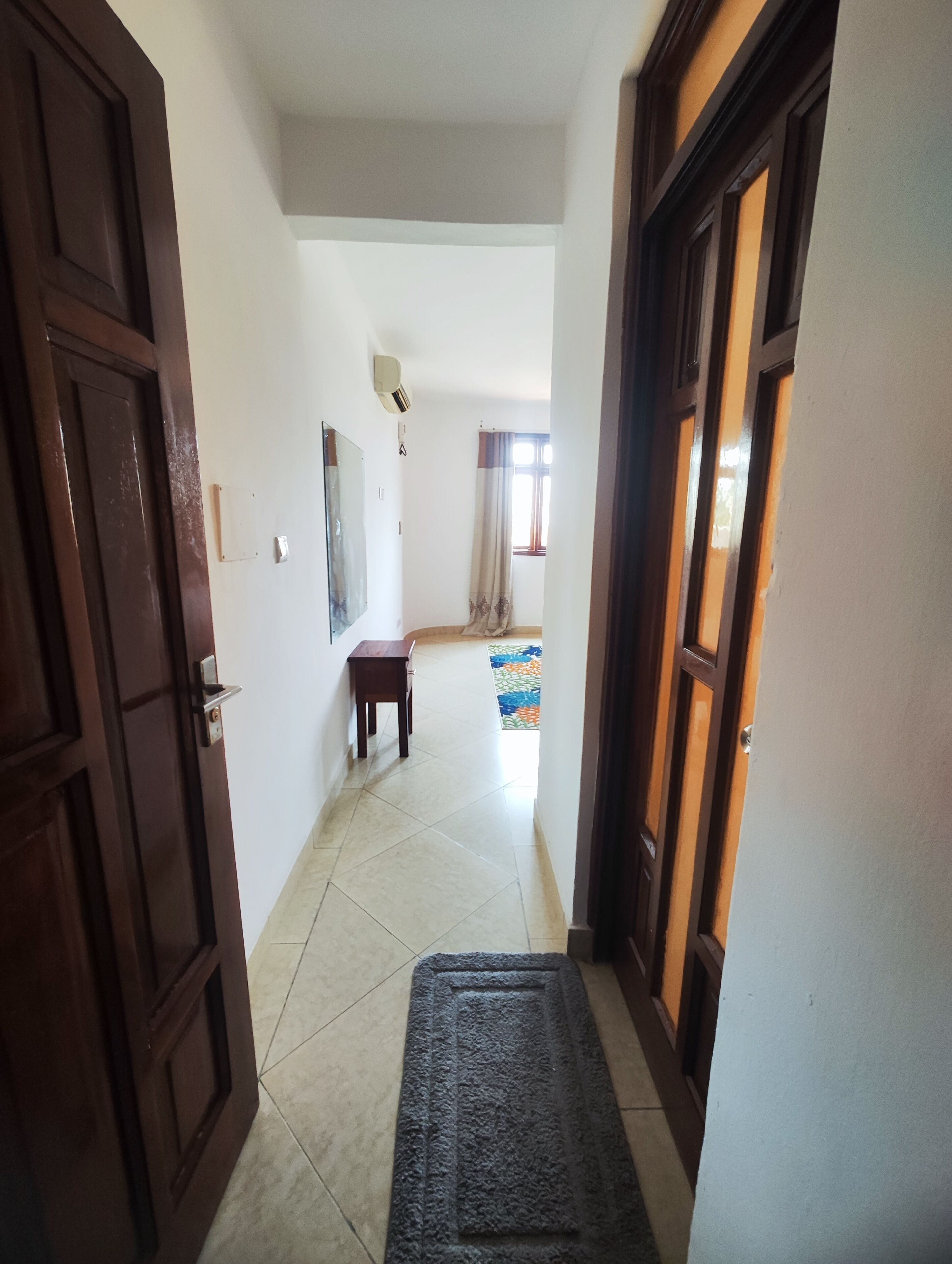 Un pasillo con una puerta de madera a la izquierda, una mesita pequeña y un tapete que conduce a una habitación con ventana y cortinas.