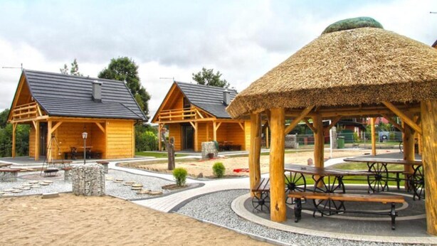 A wooden cabin-style building with a thatched roof pavilion, picnic tables, and a gravel pathway. - Holiday Homes With Fireplaces, Playground (Lubrza)