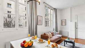 Een moderne woonkamer met een eethoek, met een bank, een kleine tafel met ontbijtartikelen en grote ramen voorzien van gordijnen.