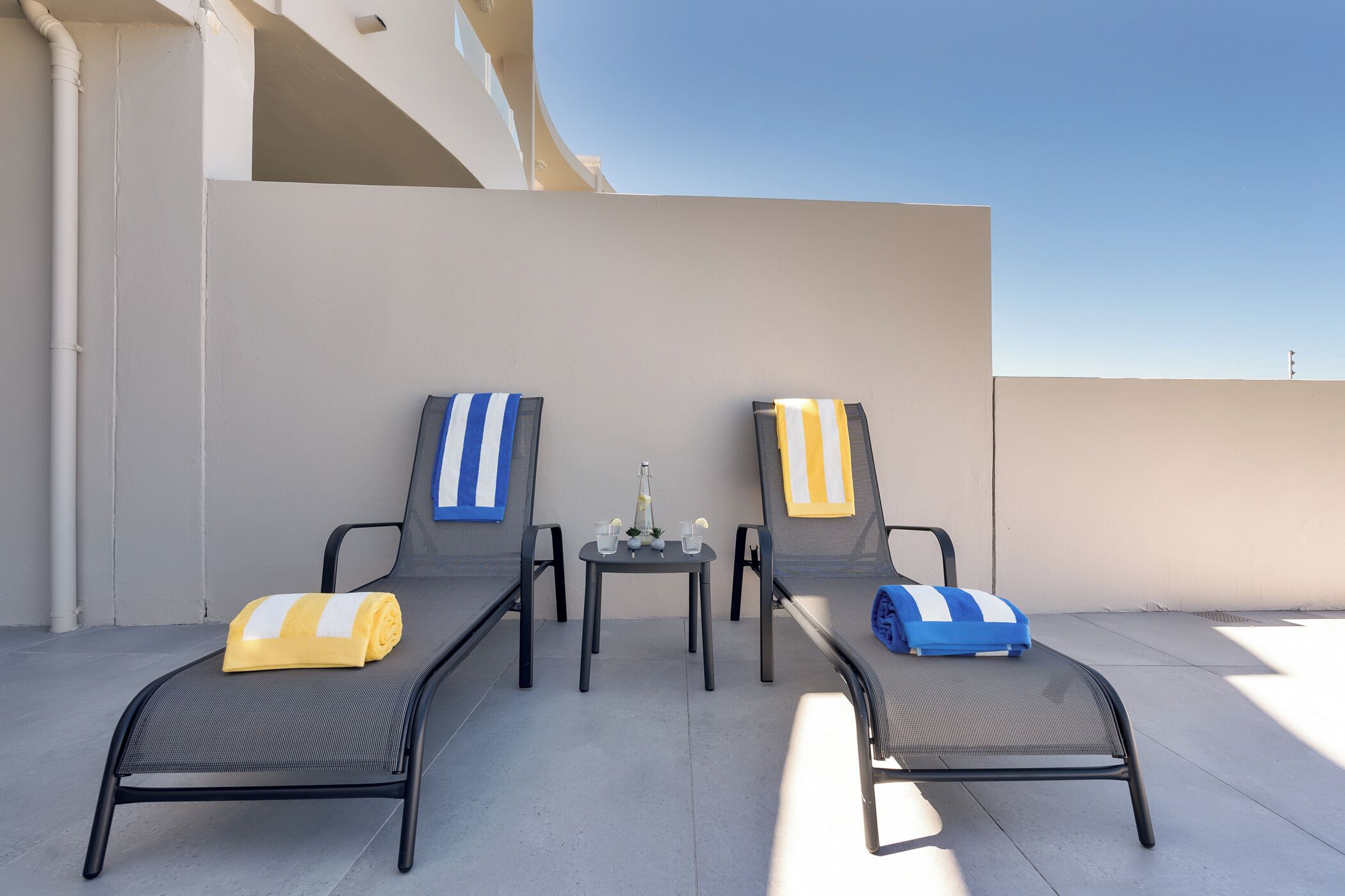 Two lounge chairs with towels, a small table with a bottle and glasses, and a clear sky.