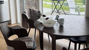 A dining area with a round table, four chairs, a bowl, and a mug. A view of a green landscape is visible through a large window.
