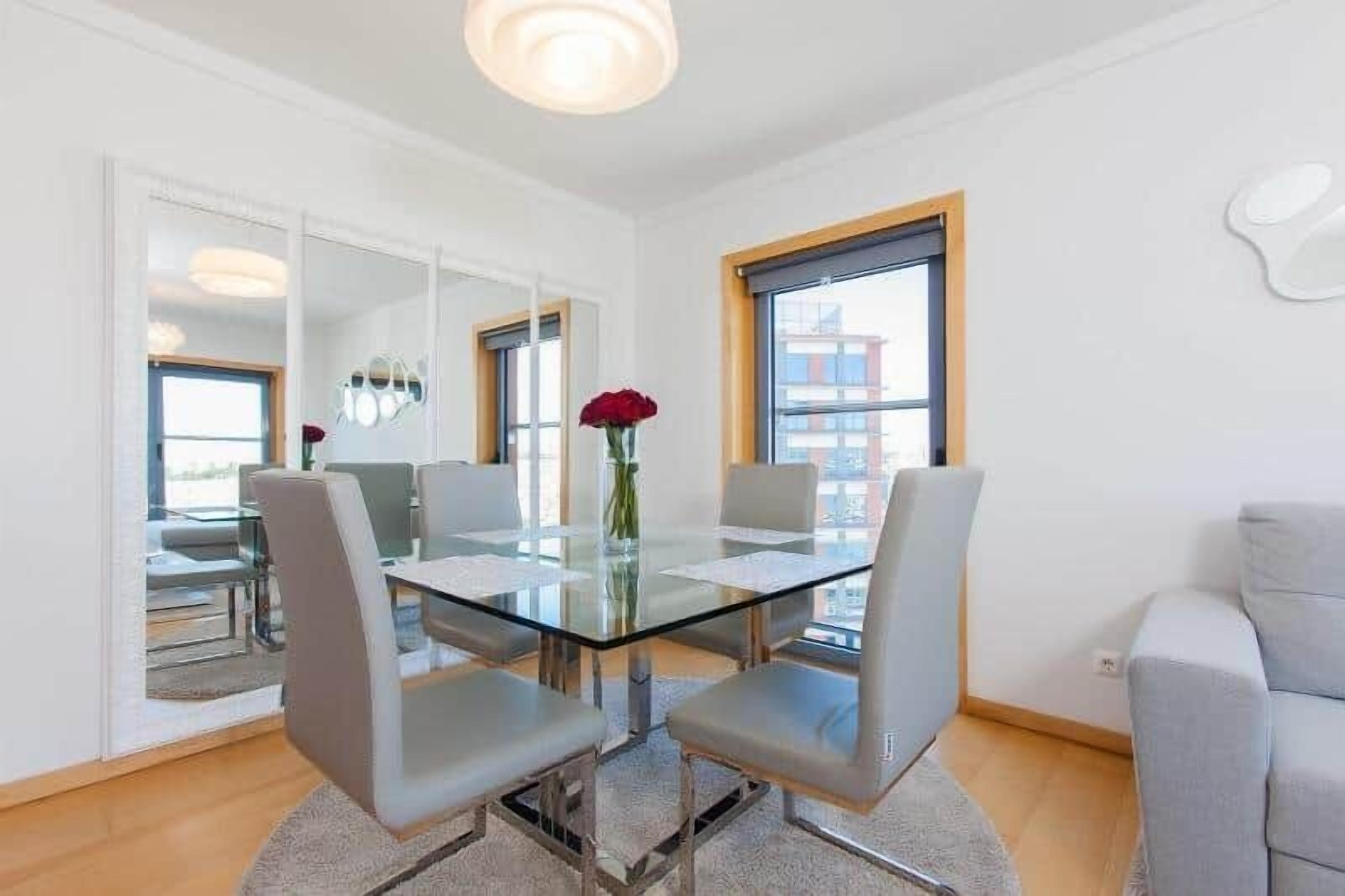 Una sala de estar moderna con una mesa de comedor de vidrio, sillas grises tapizadas, un jarrón con una flor roja y grandes ventanas que ofrecen vistas de la ciudad.