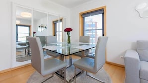 Ein modernes Wohnzimmer mit einem Esstisch mit Glasplatte, grau gepolsterten Sesseln, einer Vase mit einer roten Blume und großen Fenstern mit Blick auf die Stadt.