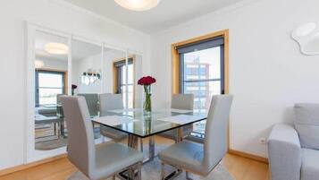 Una sala de estar moderna con una mesa de comedor de vidrio, sillas grises tapizadas, un jarrón con una flor roja y grandes ventanas que ofrecen vistas de la ciudad.