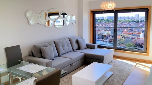 Ein modernes Wohnzimmer mit einem grauen Ecksofa, einem Glastisch und einem großen Fenster mit Blick auf die Stadt.