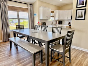 Un espace repas avec une grande table et des bancs, des meubles blancs et une vue sur l’extérieur.