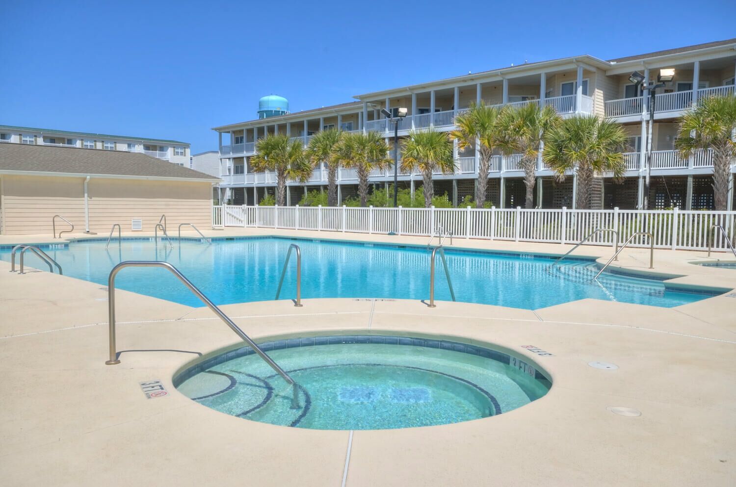 A swimming pool with a hot tub, surrounded by a white fence and palm trees, with a building in the background.