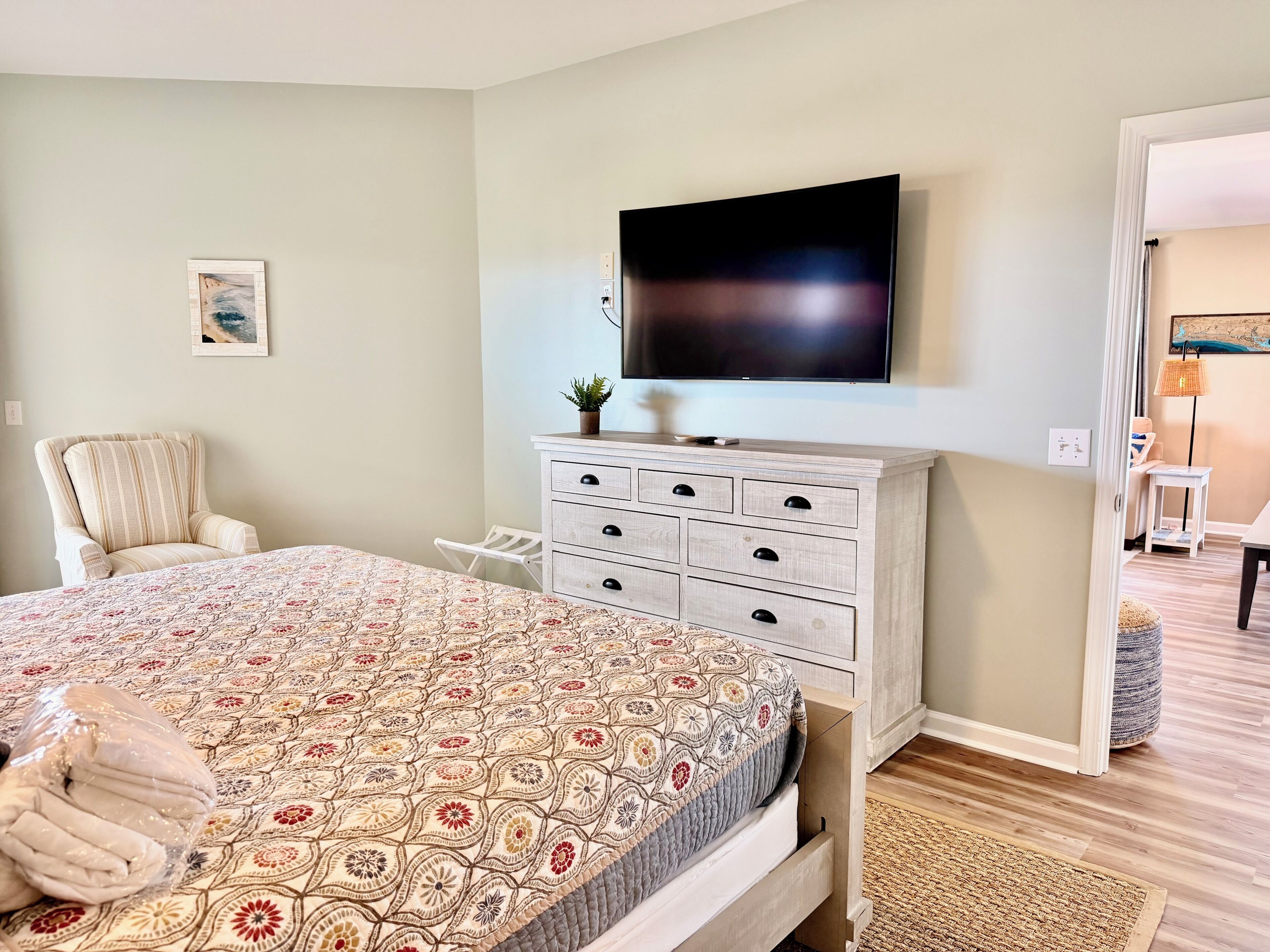 A bedroom with a bed, a television mounted on the wall, a chair, a dresser, and a small table with a lamp.