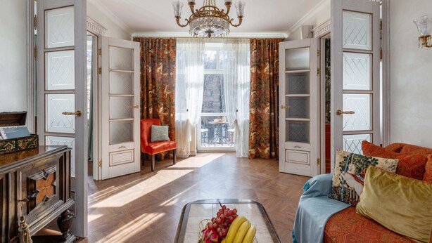 Una habitación amplia con una araña, un sofá rojo, una mesa de comedor con sillas y vistas a través de una ventana con cortinas.