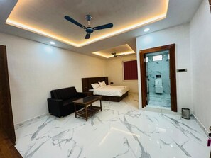 Camera d'albergo con un letto, una poltrona, un tavolino da caffè, una ventola a soffitto e un bagno visibile attraverso una porta in vetro.