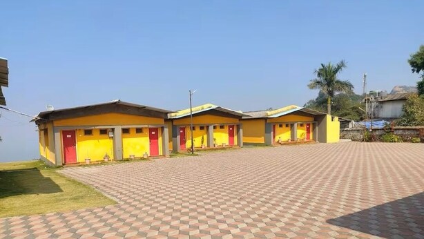 A row of yellow buildings with red doors, a paved walkway, and a palm tree in the background. - Saputara valley View (Ahwa)