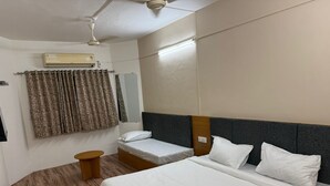 A hotel room with a large bed, a smaller bed, a wooden side table, a ceiling fan, an air conditioning unit, and a window with curtains. - Saputara valley View (Ahwa)