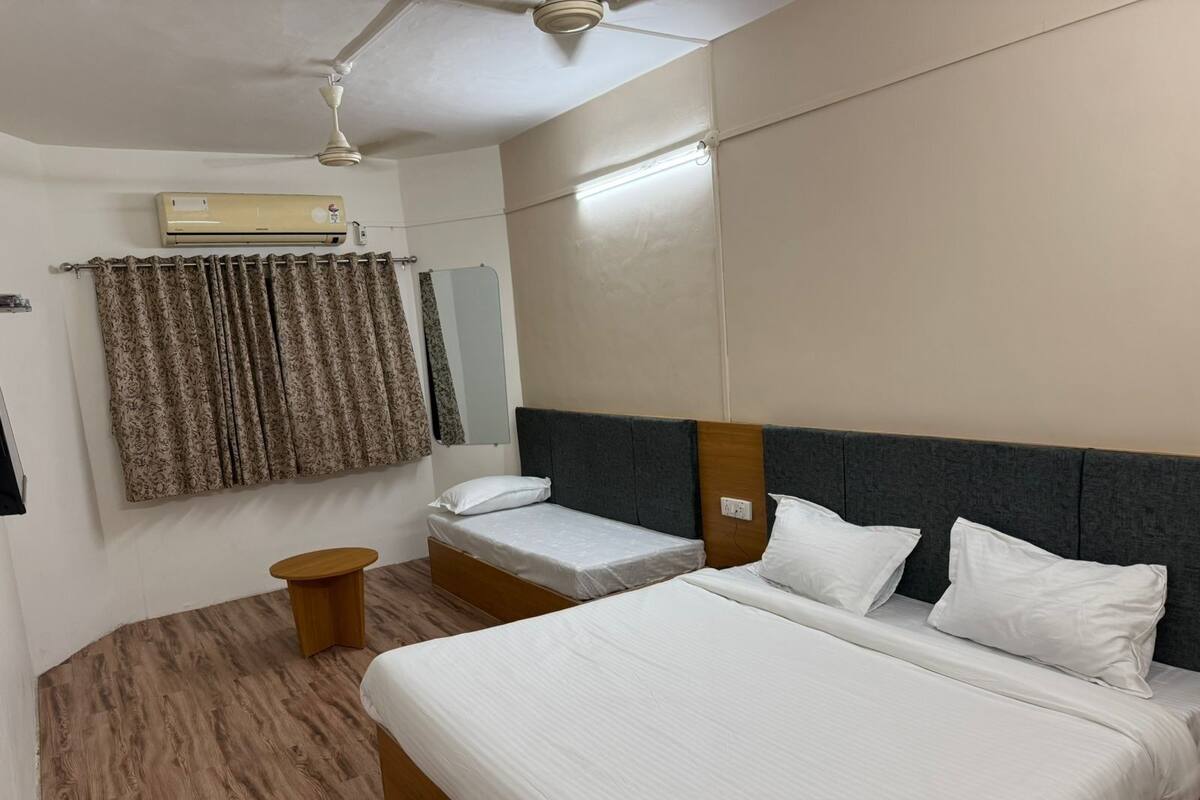 Quarto de hotel com uma cama grande, uma cama menor, uma mesinha de cabeceira de madeira, um ventilador de teto, um aparelho de ar condicionado e uma janela com cortinas.