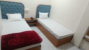 A bedroom with two beds, each with a blue headboard and white bedding. There is a nightstand with a phone and a red floral blanket on the floor. - Hotel Aadi Shree (Ujjain)