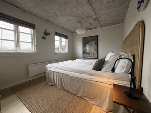 A bedroom with a bed, a bedside table, a lamp, a window, and a wall-mounted picture.