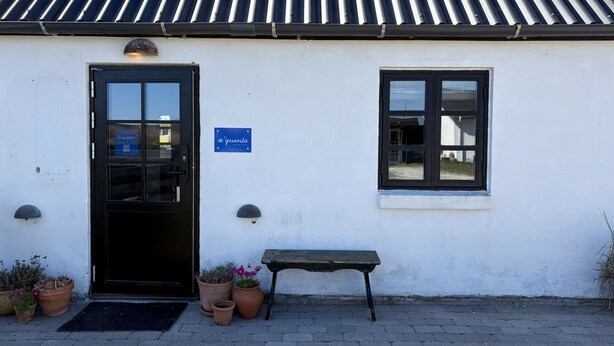 A building with a black door, a window, and a sign that reads 'a'quinta'. There are potted plants and a bench outside.