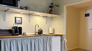 Angolo cottura con forno a microonde, macchina del caffè e lavello. Sopra il lavello, uno scaffale con bicchieri e una pianta in vaso.