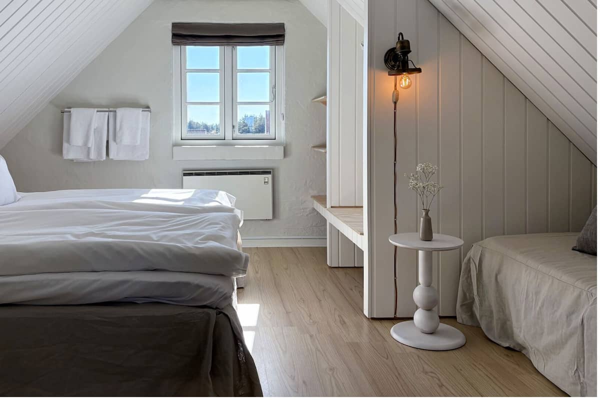 Une chambre avec un plafond en pente, une fenêtre ornée de rideaux blancs, une table de chevet avec un vase, et un lit recouvert de linge de maison blanc.