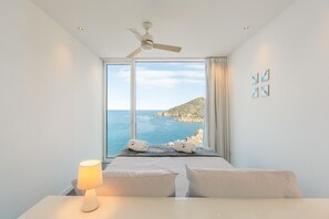 Un dormitorio con cama, lámpara de noche, ventilador de techo y vista al mar.