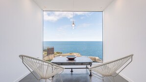 Ein modernes Zimmer mit großem Fenster mit Meerblick, zwei weiße Gestellstühle, ein Tisch mit Schüssel und eine Pendelleuchte.