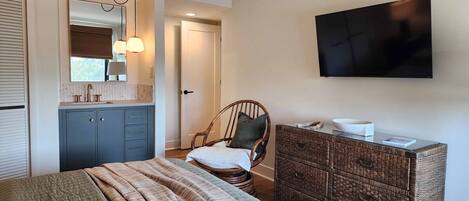 Une chambre à coucher comprenant un lit, une commode, une chaise, un téléviseur et une fenêtre avec des stores.