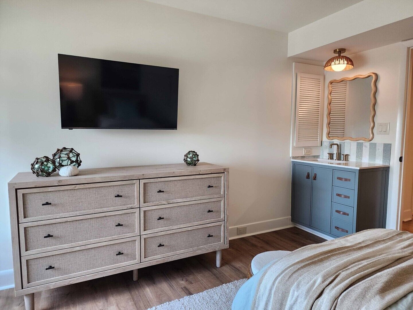 A bedroom with a wooden dresser, a television, a bathroom with a mirror and sink, and a bed with a blanket.