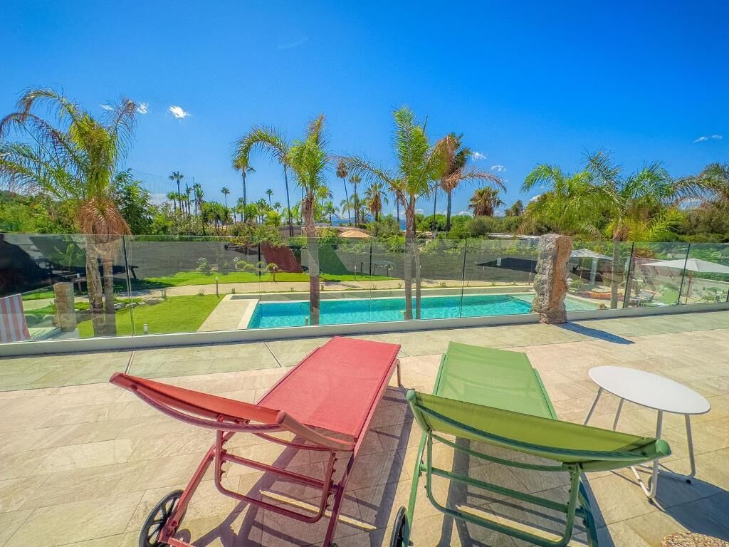 A poolside area with lounge chairs and a table, surrounded by palm trees and a clear blue sky.