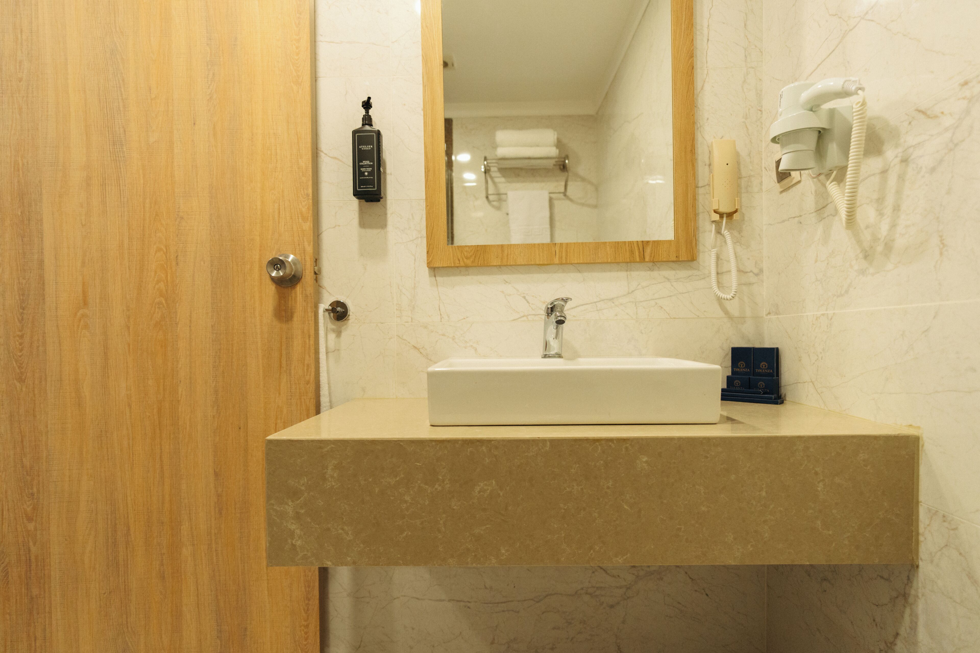 Un baño con puerta de madera, lavamanos de mármol, espejo, secador de manos y teléfono.