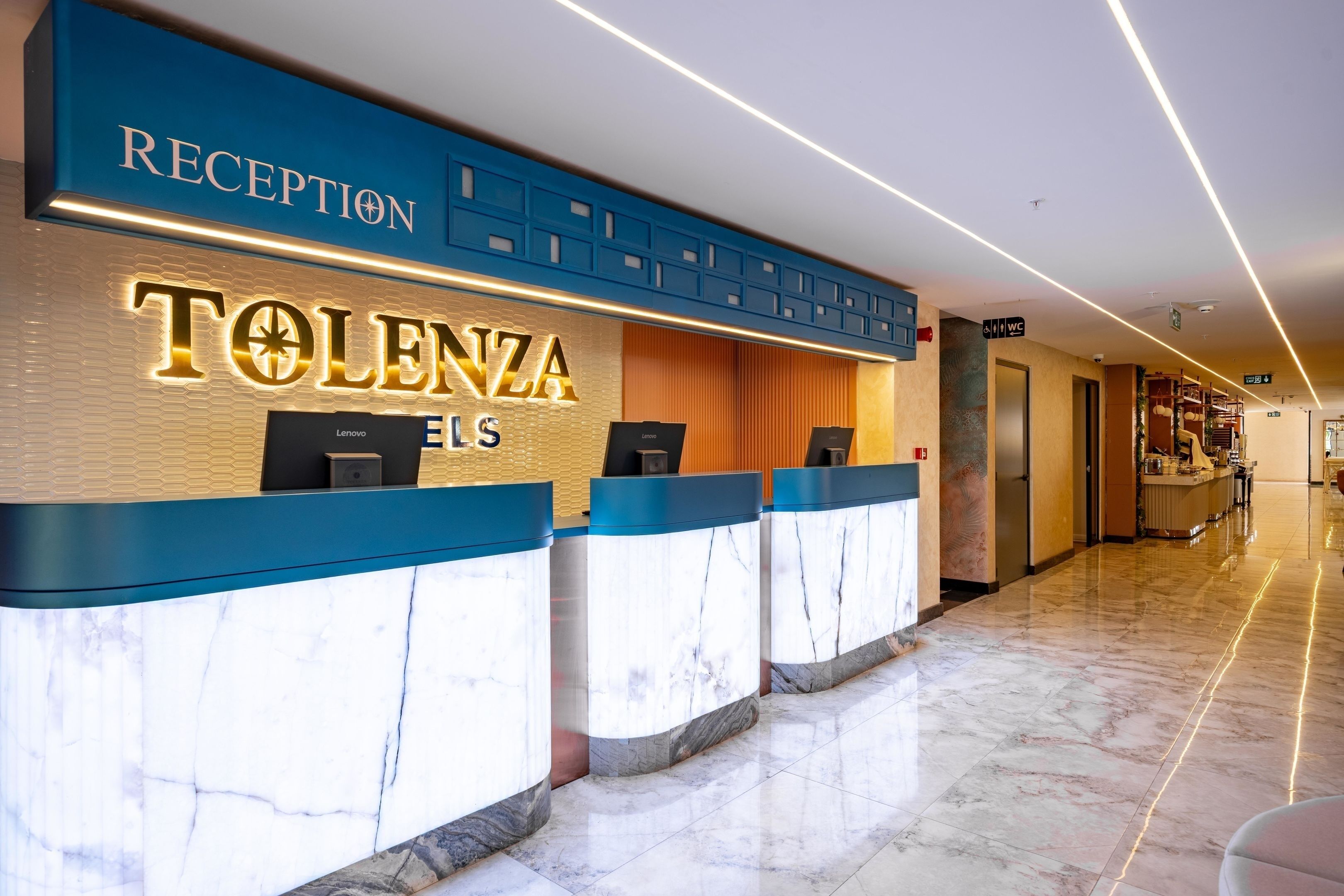 Een receptiebalie met een 'TOLENZA ELLS' bord, een receptiegedeelte en een gang naar andere delen van het hotel.