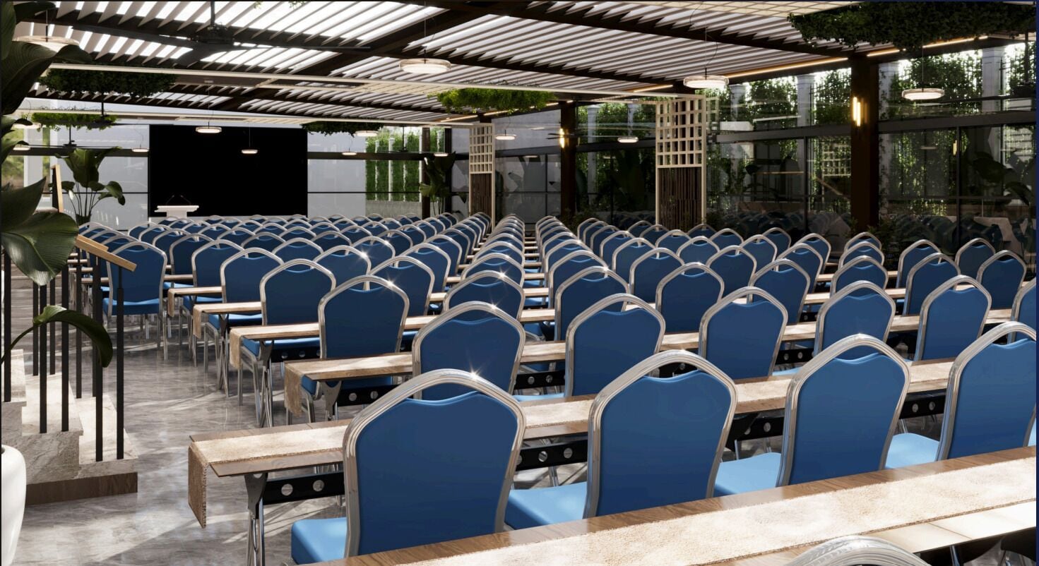 A conference room with rows of blue chairs and wooden tables, a stage, and a glass wall with greenery.