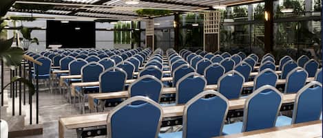 A conference room with rows of blue chairs and wooden tables, a stage, and a glass wall with greenery.