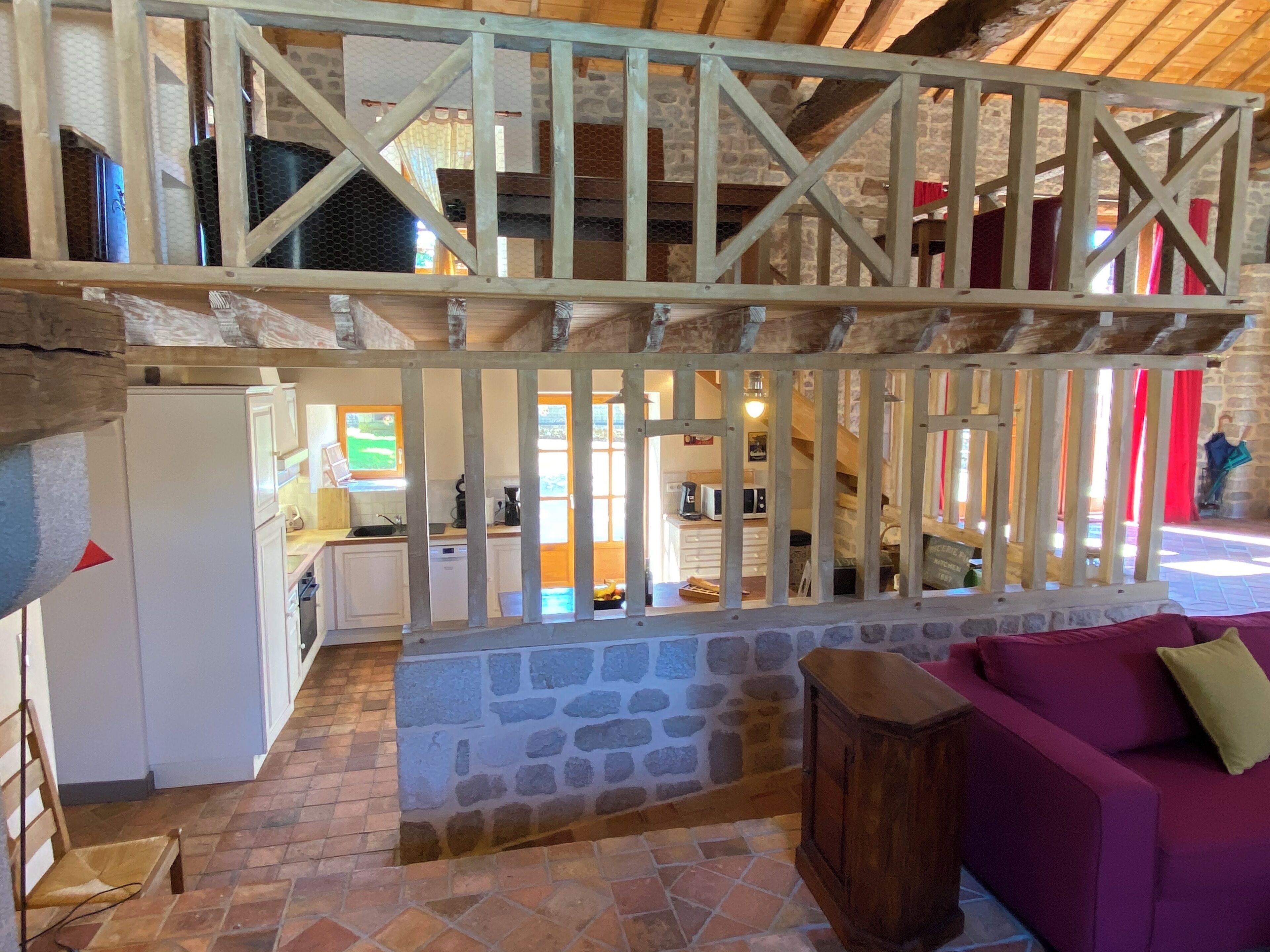 Um interior rústico com parede de pedra, vigas de madeira, um sofá roxo e uma área de cozinha visível através de um separador de madeira.