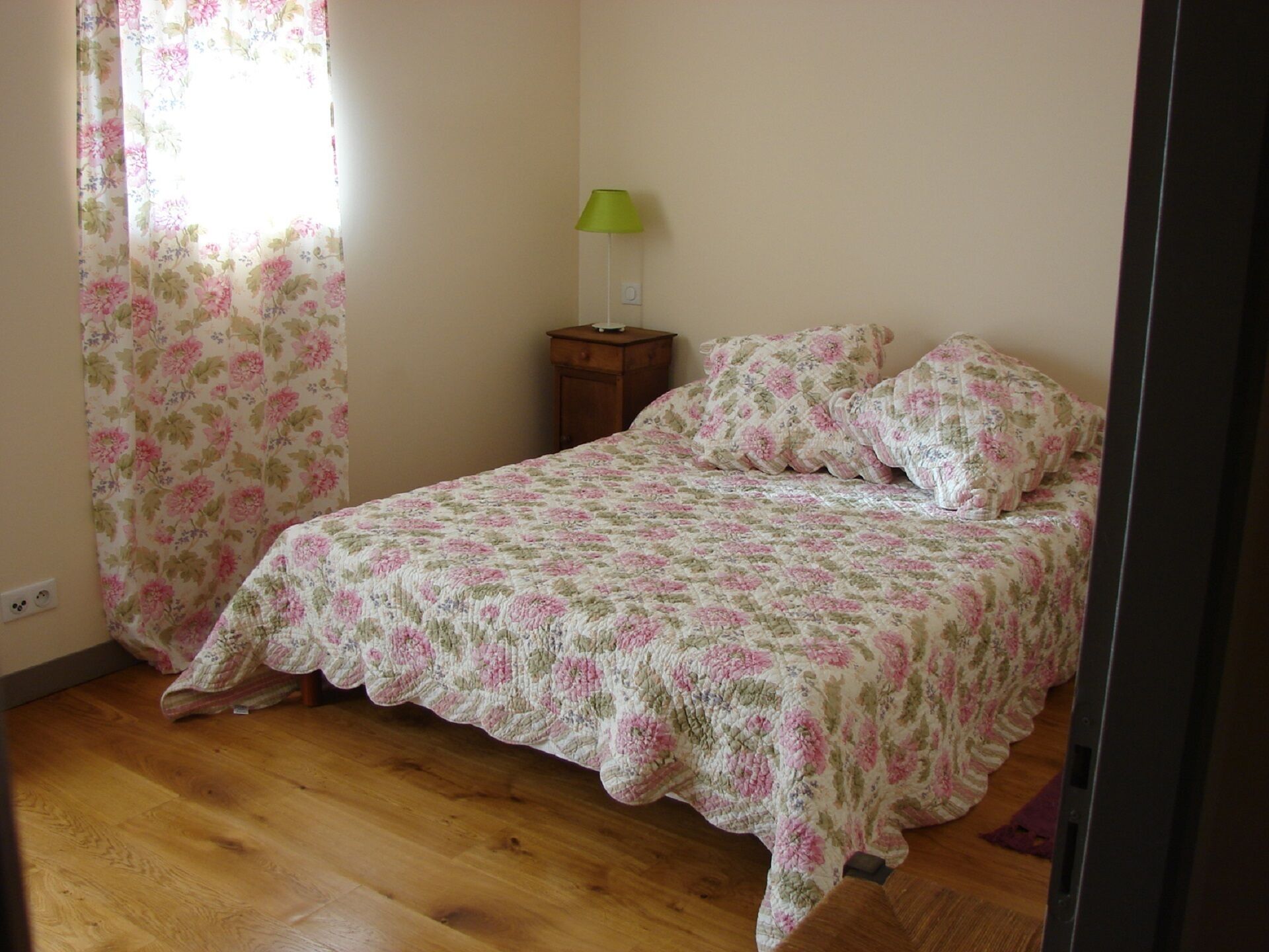 A bedroom with a floral bedspread, a wooden floor, a nightstand with a lamp, and a window with floral curtains.