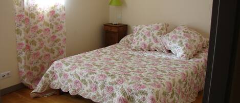 A bedroom with a floral bedspread, a wooden floor, a nightstand with a lamp, and a window with floral curtains.